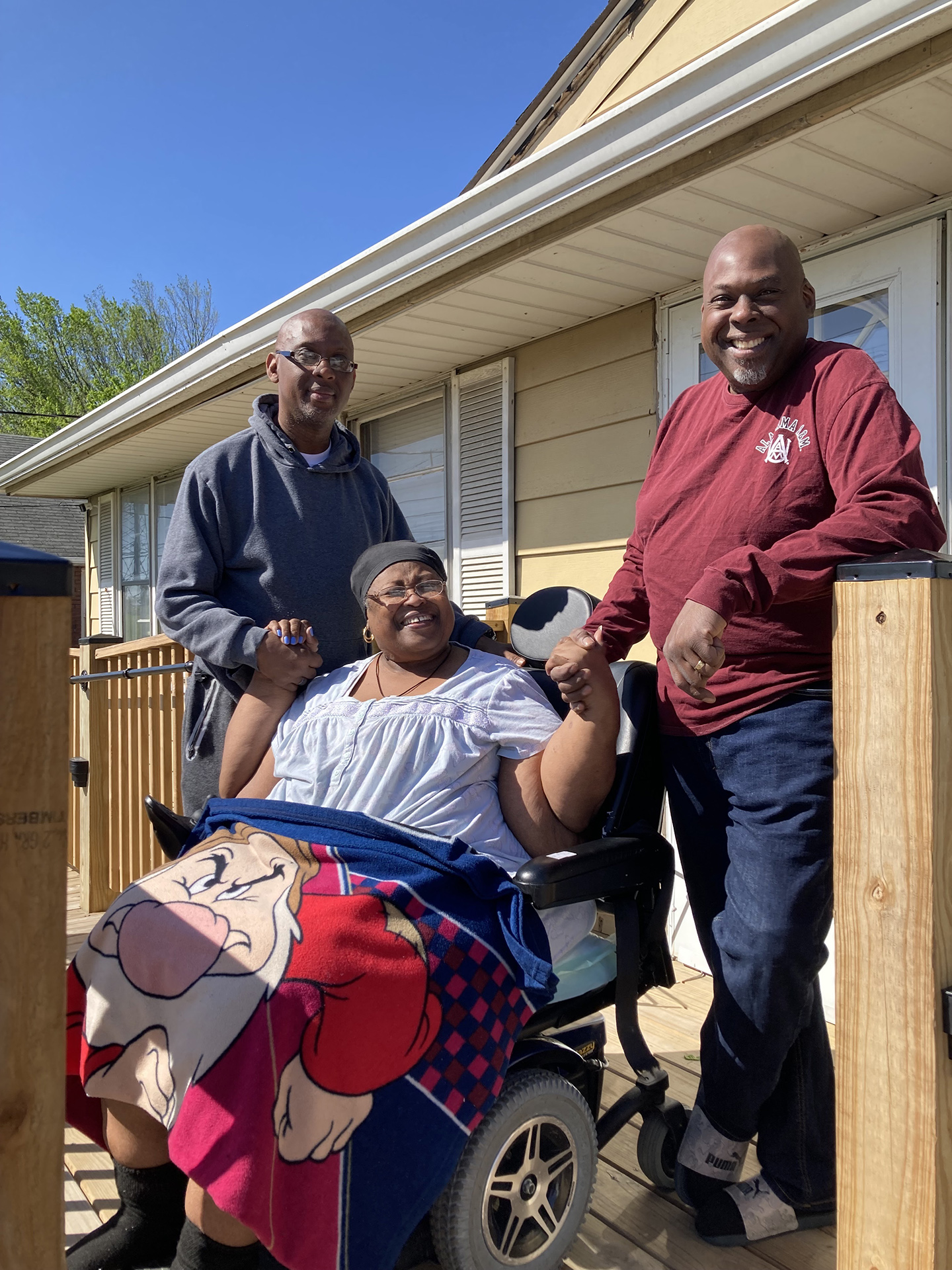 Ms. Pearlie Bowen, seated in a wheelchair, with her two sons standing beside her. They are posing on a wheelchair ramp leading up to her house.