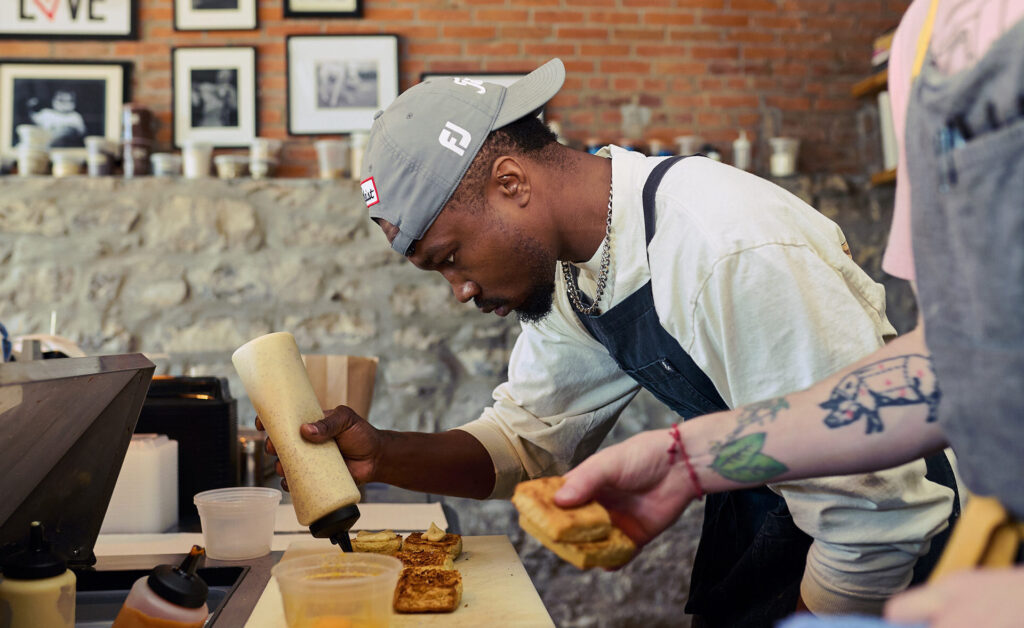 A food service worker putting sauce on pieces of toasted biscuits at a food prep counter in a restaurant.