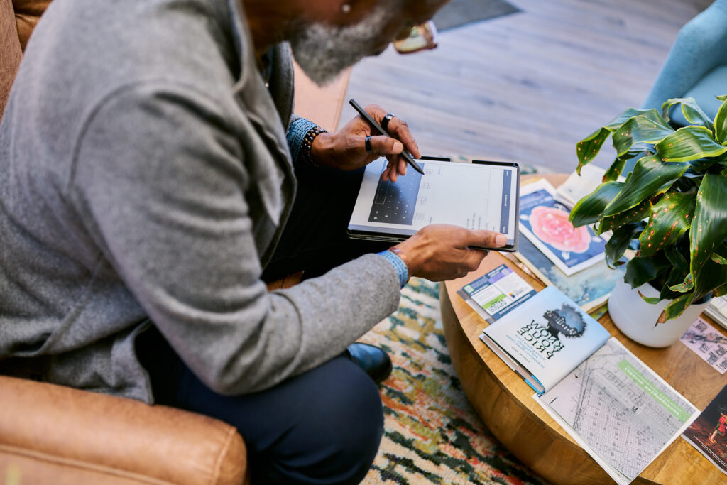A person sitting in a chair while using a tablet. A coffee table in front of the person holds a plant along with books and other reading materials.
