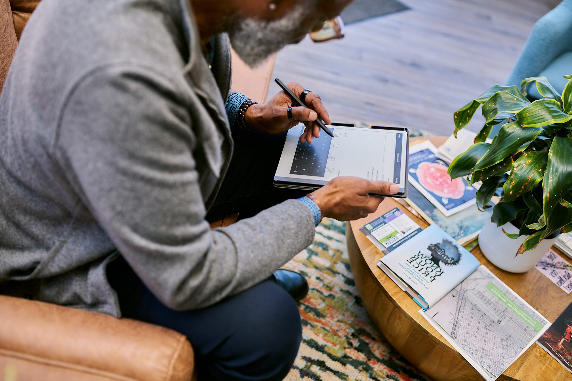 A person sitting in a chair while using a tablet. A coffee table in front of the person holds a plant along with books and other reading materials.